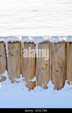 Una recinzione fatta da pali rotondi di legno intonacati e ricoperti di neve fresca profonda, retroilluminata dal sole in un giorno d'inverno. Foto Stock