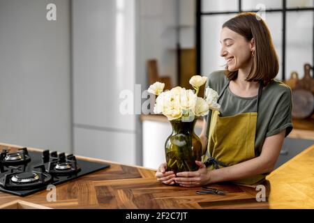 Giovane casalinga in grembiule verde decora casa con un bouquet di tulipani freschi. Fiorista con fiori in cucina a casa. Spazio di copia Foto Stock