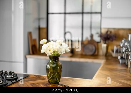 Vaso con fiori bianchi su elegante fondo interno cucina con tavolo in legno. Concetto di intimità domestica. Bouquet di tulipani in vaso di vetro verde. Foto Stock