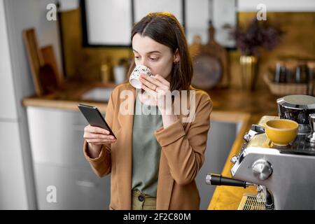 Giovane donna in giacca marrone in fretta di lavorare al mattino, bere caffè e guardare al telefono. Cucina dal design elegante. Routine quotidiana di donna d'affari. Foto Stock
