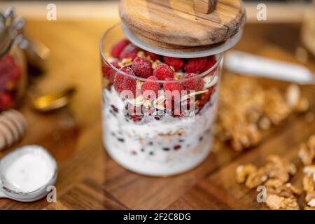 Sana colazione con muesli in vaso di vetro con lampone e yogurt biologico su tavolo in legno in cucina con noci sullo sfondo. Foto Stock