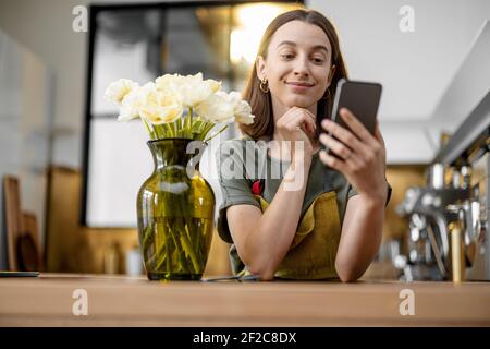 Giovane casalinga in grembiule verde ordinando con smartphone un bouquet di tulipani freschi. Fiorista con fiori in cucina a casa. Foto Stock