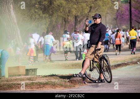 4-6-2018 Tulsa USA - uomo in bicicletta con cappello da baseball e i pantaloncini da carico e gli occhiali da sole si fermerono per scattare foto al telefono di partecipanti in holi colore correre con Foto Stock