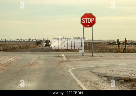 Strade di campagna - segnale di stop con grandi cavalletti a tombino all'incrocio con il pick-up con rimorchio che guida verso di esso in una nuvola di polvere Foto Stock