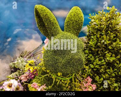 Testa di coniglio topiario verde decorativo con fiori intorno al collo e circondato da verde di fronte a sfondo blu simile al cielo - ottimo per la primavera o Foto Stock