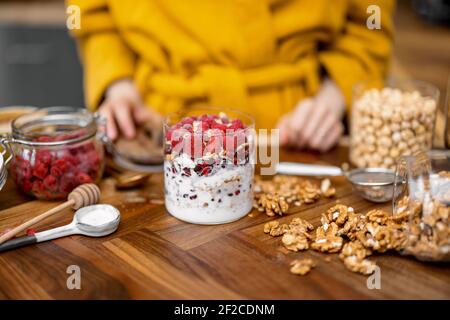 Colazione salutare con muesli in vaso di vetro con lampone e yogurt biologico su tavolo in cucina. Donna in accappatoio giallo su sfondo. Foto Stock
