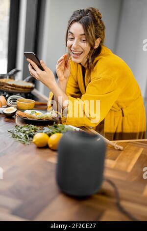 Donna felice in accappatoio che controlla i dispositivi domestici con comandi vocali, parlando a una colonna intelligente durante la colazione in cucina a casa. Concetto di casa intelligente Foto Stock