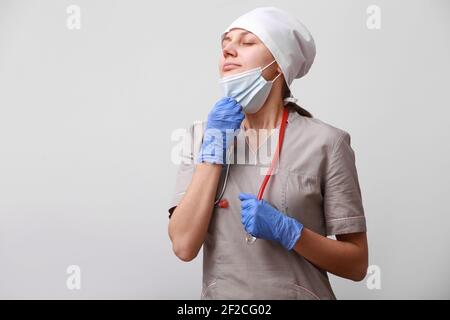 sorridente dottore con stetoscopio in uniforme. La donna sta togliendo la maschera medica protettiva dal viso per respirare aria, su sfondo bianco. Difficile Foto Stock