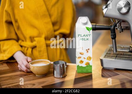 Donna in accappatoio giallo che prepara cappuccino con latte vegetale su una macchina da caffè professionale al mattino a casa. Confezionamento del latte vegetale vicino al caffè. Foto Stock