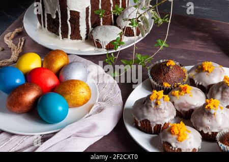 Torte pasquali fatte in casa ricoperte di glassa bianca o buccia d'arancia, colorate uova di Pasqua dipinte su piatti su sfondo di legno scuro Foto Stock