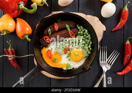 Flatlay di pasto proteico nutriente. Uova fritte, salsicce e piselli verdi in padella di ghisa su tavola di legno serviti con forchette e peperoni grezzi luminosi su b Foto Stock