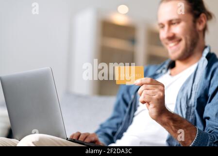 Ragazzo caucasico sorridente defocused, in abiti casual di stile, si siede sul divano a casa, tiene un laptop e una carta bancaria, effettua il pagamento online per acquisti o bollette, inserisce un numero di carta Foto Stock