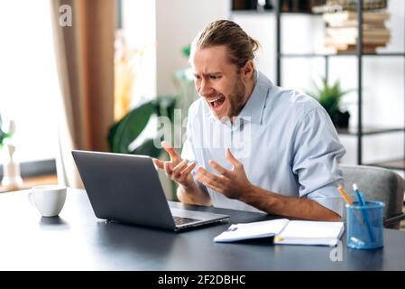 Infastidito caucasico busy business guy che guarda un portatile in confusione, ha ricevuto notizie inaspettate, ha fallito un accordo, grida, gesturing con le mani, seduto alla scrivania dell'ufficio di casa Foto Stock