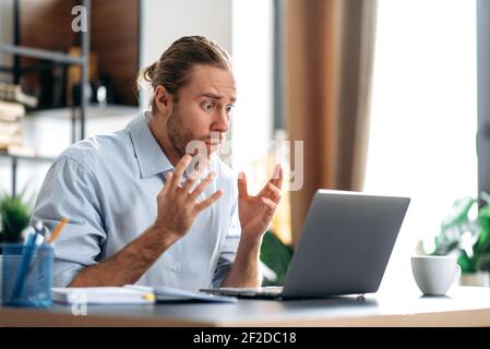 Scioccato infastidito caucasico occupato ragazzo che guarda un portatile in confusione, ha ricevuto notizie inaspettate, ha fallito un accordo, grida, gesturing con le mani, seduto alla scrivania dell'ufficio di casa Foto Stock