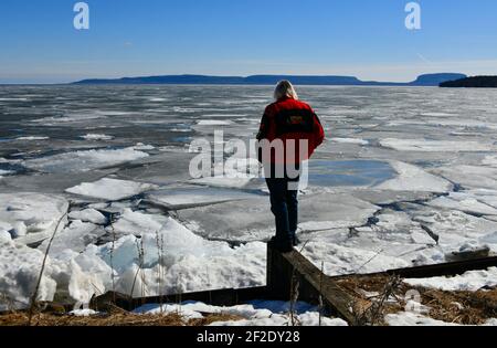 Un uomo in giacca rossa si erge a guardare la rottura del ghiaccio primaverile sul lago Superior al Chippewa Park, Thunder Bay, Ontario, Canada. Foto Stock