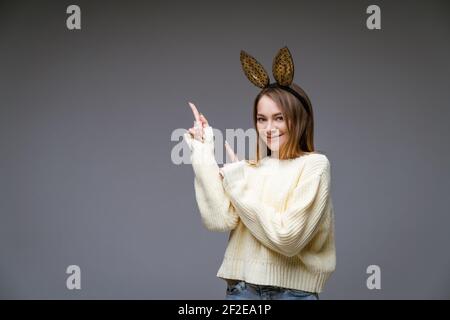 Ritratto di una bella giovane donna allegra in un maglione e nelle orecchie conigliose mostra il suo dito di lato su sfondo grigio Foto Stock