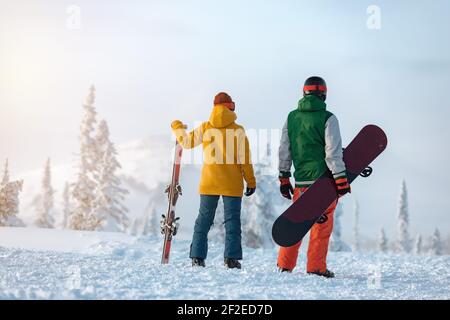 Sciatore femminile e snowboarder maschile sono in piedi sullo sfondo della montagna innevata al tramonto. Concetto di stazione sciistica Foto Stock