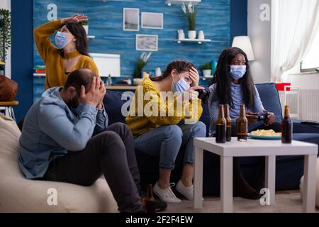 Deluso gruppo diverso di persone che giocano video giochi rispettando la divaricazione sociale a causa di orona focolaio indossare maschera viso non per diffondere il virus e si ammalano. Bottiglia di birra e patatine. Foto Stock
