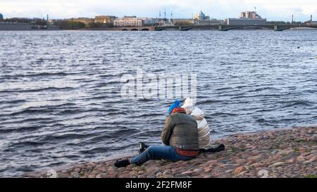 Due persone in abiti caldi sono seduti sui ciottoli della riva del fiume, vista posteriore Foto Stock