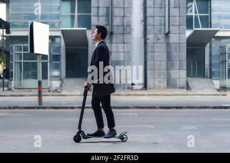 Uomo d'affari asiatico che guida uno scooter elettrico sulle strade della città per andare a lavorare al mattino. Spostamenti quotidiani che riflettono meglio il mondo del lavoro di to Foto Stock