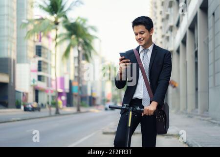 L'uomo d'affari asiatico sta guidando uno scooter elettrico e usando il suo telefono cellulare per aprire una mappa in applicazione per controllare le strade della città per lavorare nel m Foto Stock