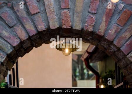la lampada da strada retrò si illumina con una luce calda sull'arco in mattoni della volta dell'antico edificio, l'architettura delle strade turistiche europee con lanterna in ferro e gl Foto Stock