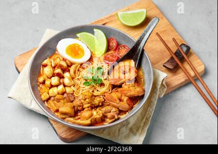 Laksa in ciotola grigia sul piano del tavolo in cemento. Zuppa di noodle al cocco con carne di pollo e gamberi di cucina peranakan. Cibo asiatico. Foto Stock