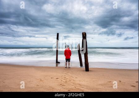 Uomo in piedi sulla spiaggia e grande onda che colpisce decadimento ponte di legno in tempo tempestoso a Pilai Beach, Phang Nga, Thailandia Foto Stock