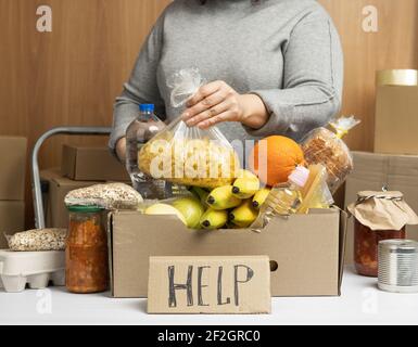 donna in un maglione grigio mette in una scatola di cartone vari alimenti, frutta, pasta, olio di girasole in una bottiglia di plastica e confetture. Donazione e volontariato Foto Stock