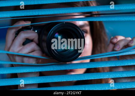 Giovane paparazzi donna con fotocamera in mano guarda fuori il finestrino attraverso le tendine Foto Stock