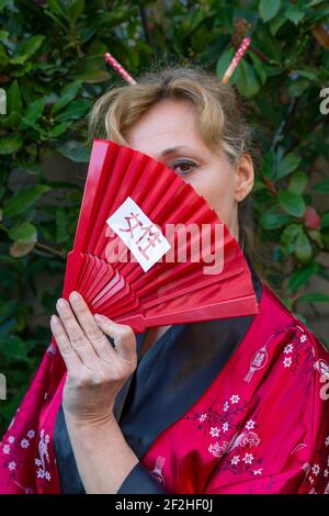 Donna bionda in abiti orientali copre il suo viso con un ventilatore rosso della mano su cui è scritta la parola donna In giapponese Foto Stock