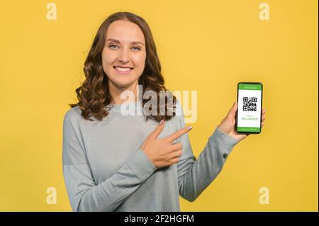 Sorridente giovane donna punta al passaporto covid on-line sullo smartphone, isolato in giallo, signora mostra un e-documento di conferma della vaccinazione, app necessaria per viaggiare durante e dopo il periodo pandemico Foto Stock