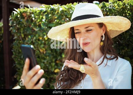 donna sorridente di 40 anni in camicia bianca con cappello che parla videochiamata su uno smartphone all'aperto vicino a parete verde. Foto Stock