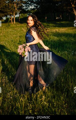 Concetto da favola per la cerimonia di laurea. Bella giovane donna brunetta in abito blu con bouquet lilla su sfondo naturale. All'aperto romantico Foto Stock