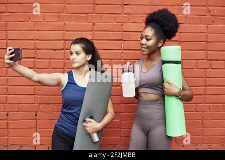 Immagine di donne atletiche giovani sorprendenti che prendono un selfie contro un muro di mattoni. Le donne nere sorridenti trascorrono il tempo insieme dopo l'allenamento sportivo. Foto Stock