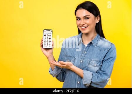 Kiev, Ucraina - 28 febbraio 2021. Concetto di clubhouse. Caucasica bella giovane donna sorridente che tiene il cellulare con l'app clubhouse raffigurata sullo schermo, in piedi contro lo sfondo giallo isolato Foto Stock