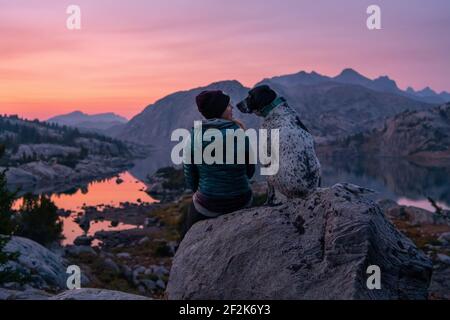 Vista posteriore della giovane donna seduta sulla roccia con il cane in montagna durante il tramonto Foto Stock