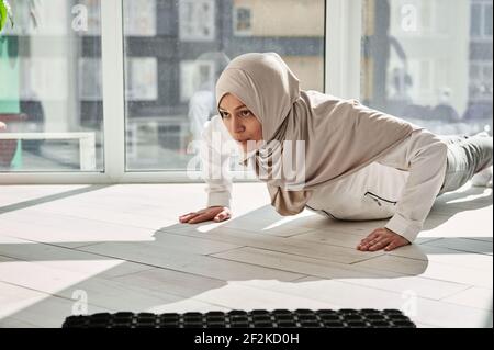 Determinata donna musulmana araba che fa push-up sullo sfondo di grandi finestre a casa in una bella giornata di sole. Concetto di allenamento domestico Foto Stock