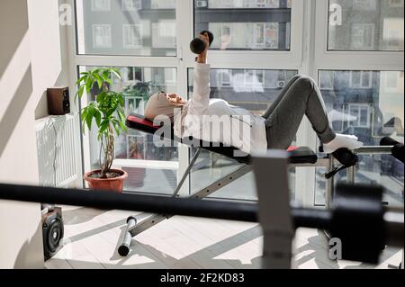 Vista laterale di una determinata donna musulmana araba con testa coperta nel hijab facendo pressa della panca del dumbbell alla palestra domestica contro lo sfondo di grandi finestre Foto Stock