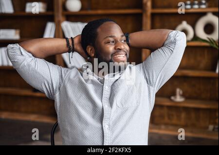 Spensierato e riuscito uomo d'affari afroamericano che prende una pausa sul posto di lavoro, un ragazzo nero in camicia casual intelligente appoggiato sulla sedia, messo le mani dietro la testa, sorride Foto Stock