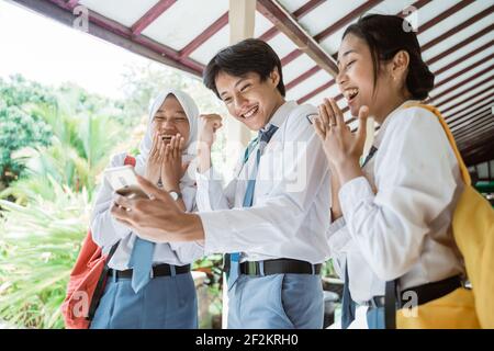 Tre studenti indonesiani delle scuole superiori guardano lo schermo usando uno smartphone per vedere l'annuncio della laurea Foto Stock