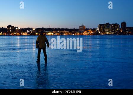 Uomo che guarda la luce della città riflessi di Barrie Canada su ghiaccio Di Frozen Kempenfelt Bay al crepuscolo blu d'ora in inverno Foto Stock