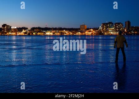 Uomo in piedi su ghiaccio spesso di Kempenfelt Bay ghiacciato a. Crepuscolo di Blue hour in inverno con le luci del centro di Barrie Ontario, Canada Foto Stock