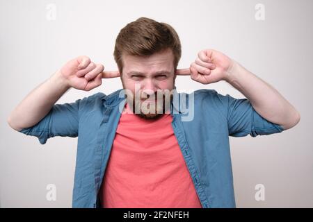 Uomo caucasico dispiaciuto e irritato stanco di rumore forte, coprendo le orecchie con le dita e accigliando alla macchina fotografica. Sfondo bianco. Foto Stock