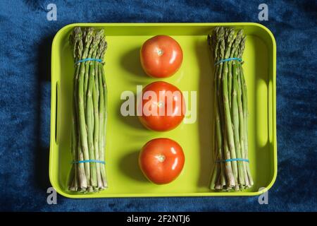 Due grappoli di asparagi con 3 pomodori a composizione geometrica su un vassoio verde colorato e fondo tessuto blu. Verdure e frutta. Foto Stock