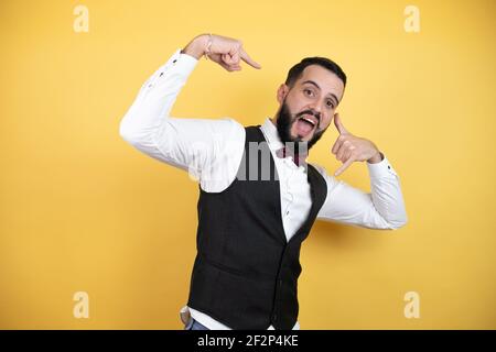 Giovane uomo con barba con cravatta ad arco e gilet che fa il gesto "chiamami" con le mani. Foto Stock