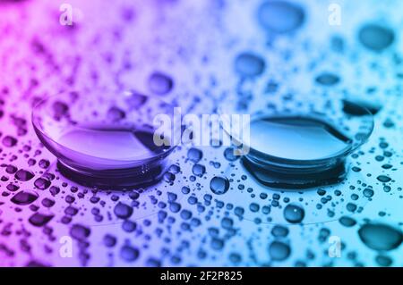 Lenti a contatto, su sfondo rosa-blu-verde. Due lenti a contatto con gocce d'acqua su sfondo rosa-blu. Foto Stock