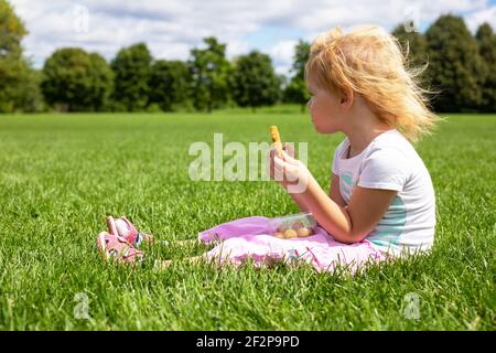 Bambino seduto sull'erba nel prato e con uno spuntino in una giornata estiva soleggiata. Foto Stock