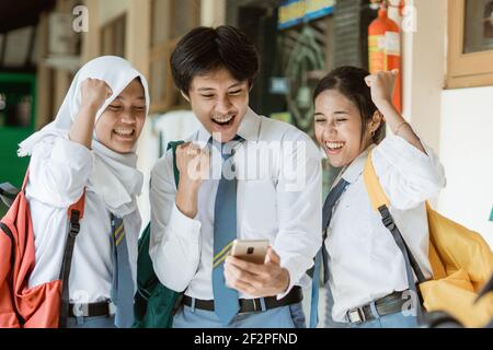 primo piano di tre studenti in uniforme di scuola superiore con entusiasmo utilizzare uno smartphone per visualizzare gli annunci congiunti Foto Stock