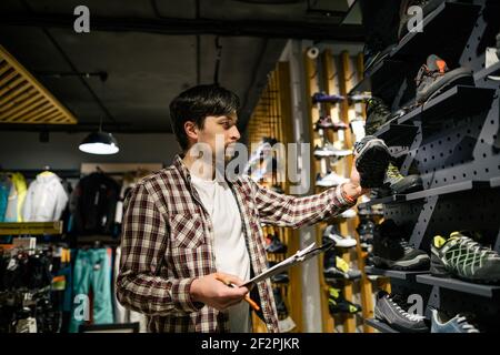Il proprietario di affari in negozio di sport lavora con vetrina trekking e scarpe da montagna per la vendita, tenendo gli appunti in mano, controlla la disponibilità di Foto Stock